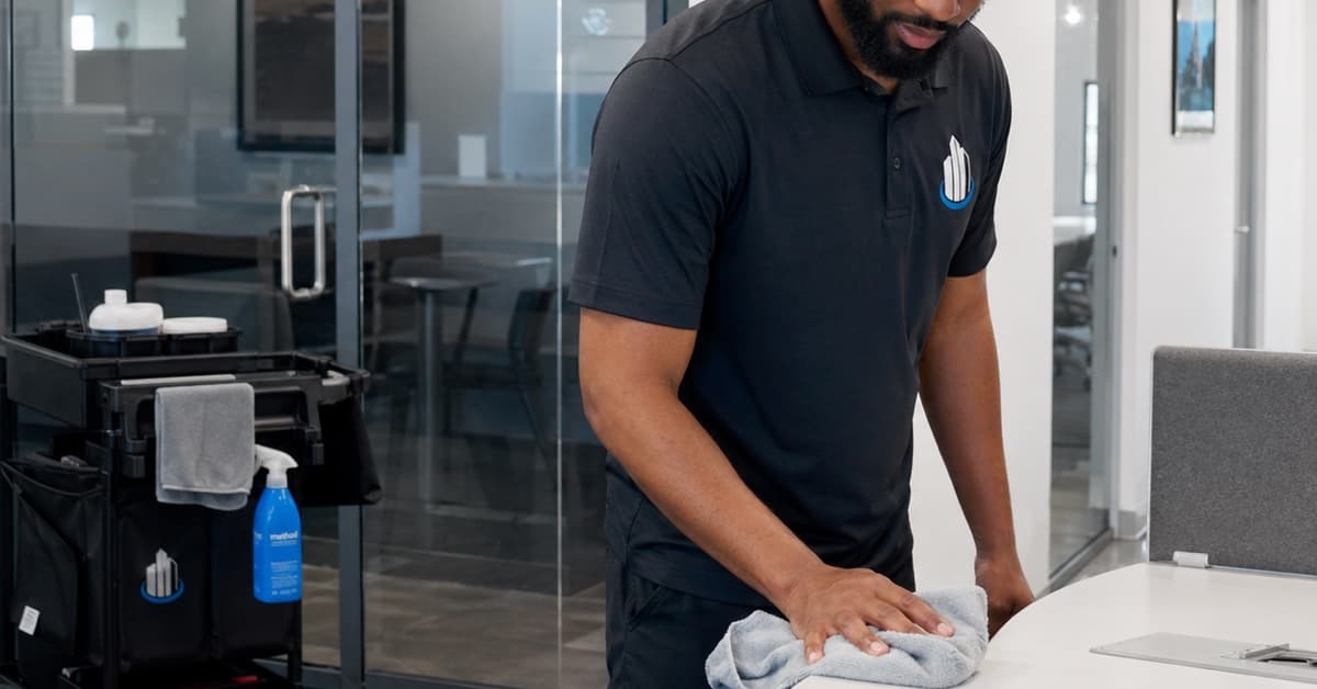 Uniformed commercial cleaner wiping an office table with a janitorial cart nearby